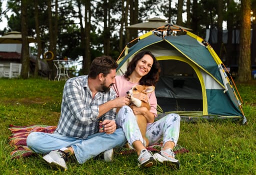 Young married couple with their corgi dog sitting in the forest near tent and looking at sunsetYoung married couple with their corgi dog sitting in the forest near tent and looking at sunset