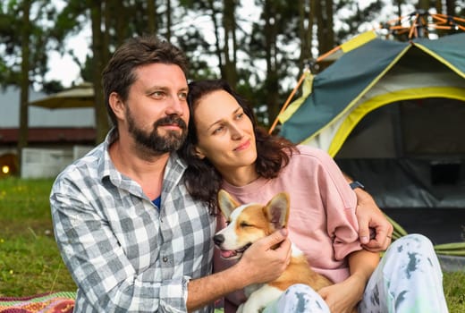Young married couple with their corgi dog sitting in the forest near tent and looking at sunset