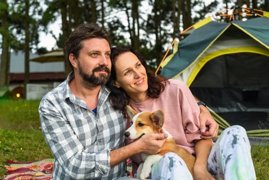 Young married couple with their corgi dog sitting in the forest near tent and looking at sunset