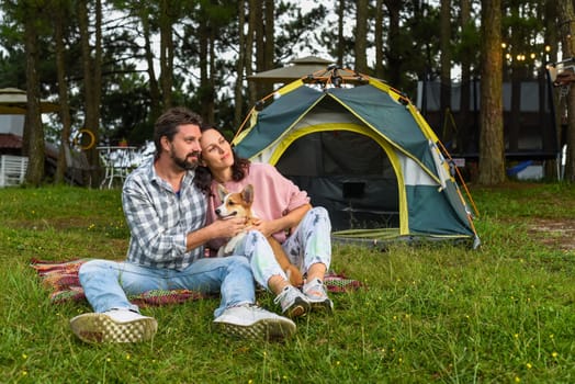 Young married couple with their corgi dog sitting in the forest near tent and looking at sunset