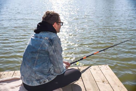 Young fisherman in denim jacket sitting on wooden pier, fishing in the lake. he holding the rod and looking at float.