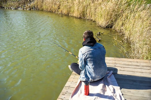 Young fisherman in denim jacket sitting on wooden pier, fishing in the lake. he holding the rod and looking at float.