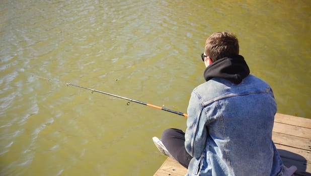 Young fisherman in denim jacket sitting on wooden pier, fishing in the lake. he holding the rod and looking at float.