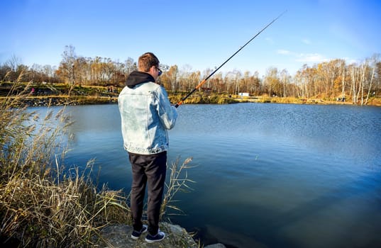 Young fisherman in denim jacket standing on stone, fishing in the lake. he holding the rod and looking at float against autumn forest in park.