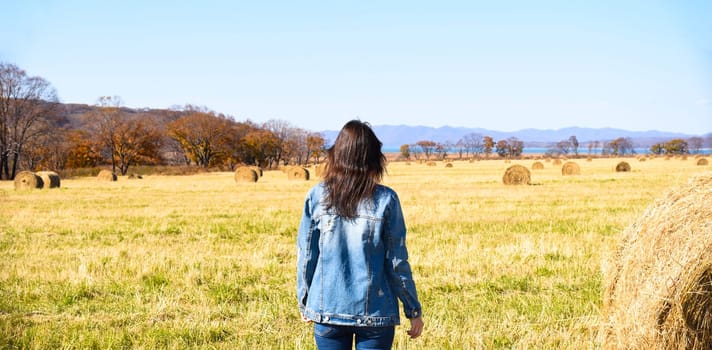 Young brunette woman in denim jacket standing near haystack in field with bales of hay. she looking at mountains and the autumn forest.