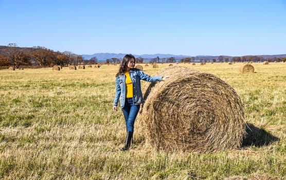 Young brunette woman in denim jacket standing near haystack in field with bales of hay