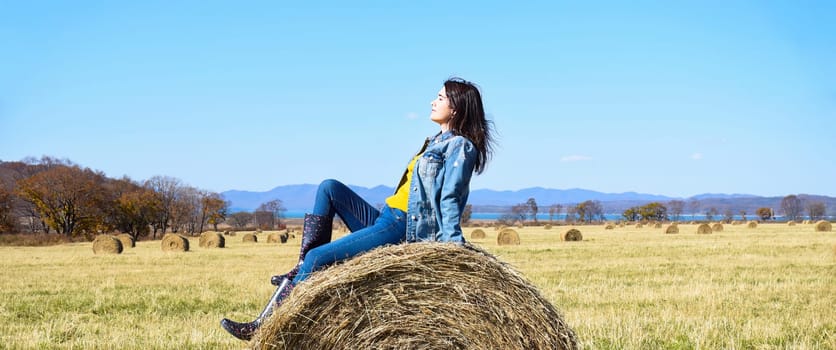 Young brunette woman sitting on top of haystack in field. she squinting from the sun