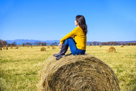Young brunette woman in yellow sweater and rubber boots sitting on top of haystack in field. she looking at mountains, Japanese sea and the autumn forest. Primorsky kray, Russia.