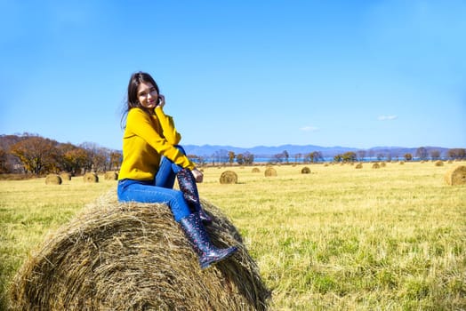 Young brunette woman in yellow sweater and rubber boots sitting on top of haystack in field