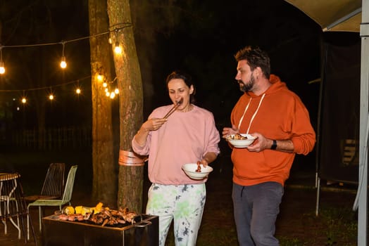 Married couple man and woman eating barbecue sausages at night near lightning garlands in the backyard