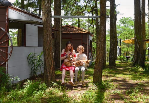 two sisters ride together on a swing and hold their corgi dogs in park