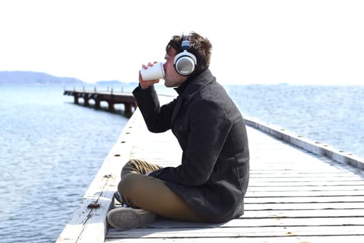 Young man listening to music and drinking coffee, sitting alone on pier on a sunny day