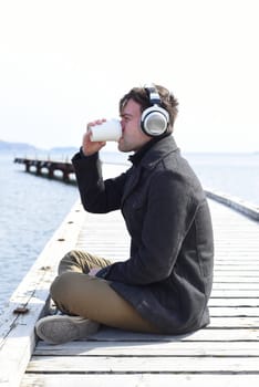 Young man listening to music and drinking coffee, sitting alone on pier on a sunny day