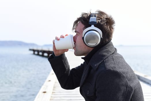 Young man listening to music and drinking coffee, sitting alone on pier on a sunny day