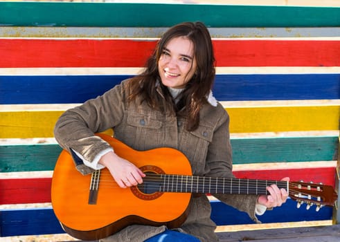 Young woman playing guitar and laughing, sitting on a bench against colorful background
