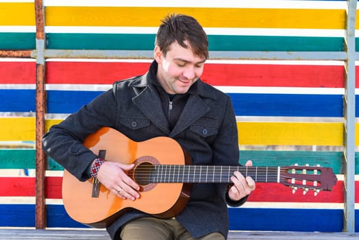 Smiling young man playing guitar and sitting against colorful wall