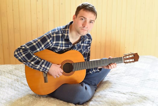 Young man playing guitar and sitting on bed on weekend morning. looking at the camera