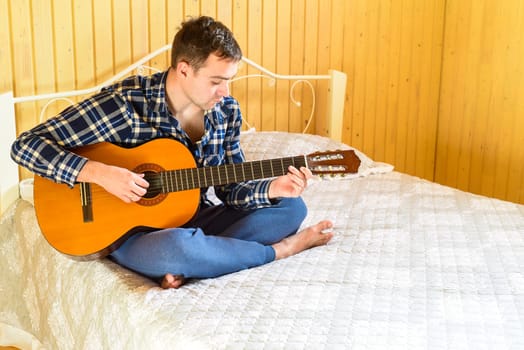 Young man playing guitar and sitting on bed on weekend morning. looking at the camera