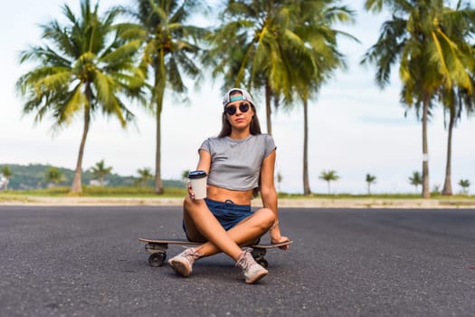 Young beautiful woman in a cap sitting on a skateboard and drinking coffee
