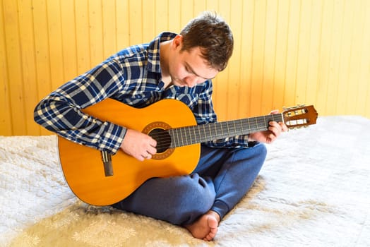 Young man playing guitar and sitting on bed on weekend morning. looking at the camera