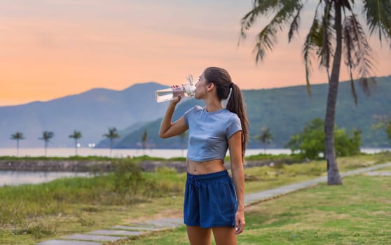 Young slim woman drinking water after exercises and sport against sunset and beach with palms