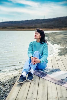 49 year old Russian woman in sweater sitting on a pier and thinking with eyes closed