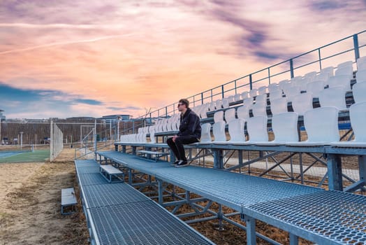 One young man sitting on empty football tribune on the street under sunset