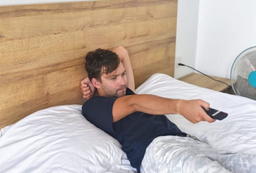 Man watching TV  and holding a TV remote on a bed with fan