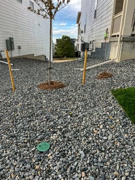 Castle Rock, Colorado, USA-May 17, 2023-A side yard between two houses covered in gray gravel with young trees recently planted and supported by yellow stakes The area is landscaped with mulch around the tree bases and a few utility markers on the ground The house on the right has a small porch with stairs while both homes have exterior utilities visible along their walls The sky is partly cloudy suggesting an outdoor improvement project in progress