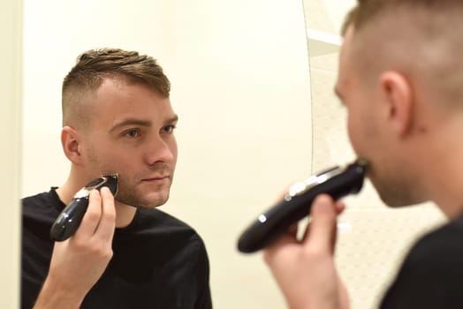 Young man shaving with trimmer and looking in the mirror in a bathroom