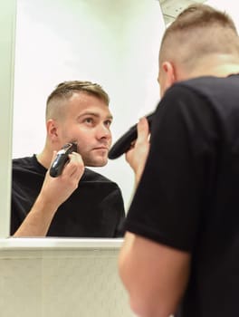 Young man shaving with trimmer and looking in the mirror in a bathroom