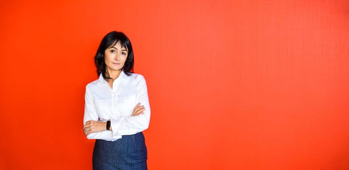 45 y.o. Russian brunette woman in white shirt with crossed arms looking at the camera and standing against red background. Success and business concept. Copy space