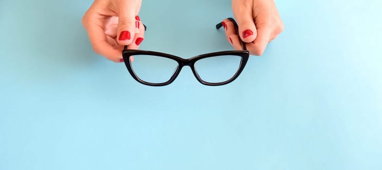 Female hands with red nail polish holding black glasses for eye vision with frame Cat's eye on a blue background. Copy space