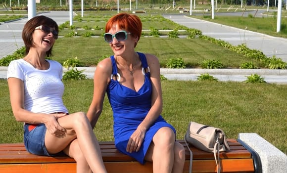 Two young russian sisters in sunglasses sitting on the bench and sincerely laughing together on a summer day.