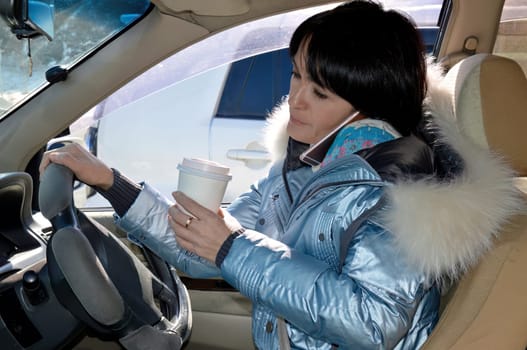 Young attractive woman in jacket driving the car, drinking coffee and talking on phone on a winter day