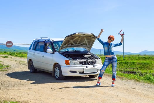 Red-haired woman with a screwdriver and pliers on high heels standing near a broken car with open hood on a country road and barrier with stop sign