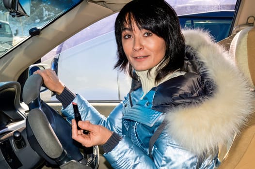 Young woman driving the car and make up lips with lipstick. She smiling with teeth and looking at the camera