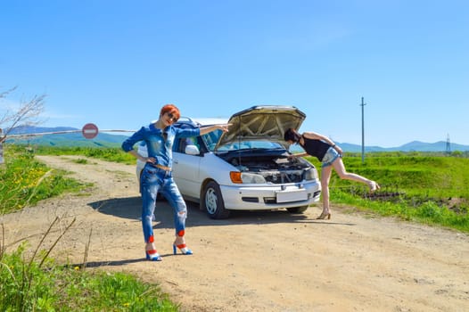 Red-haired woman with a screwdriver and pliers on high heels standing near a broken car with open hood on a country road and barrier with stop sign