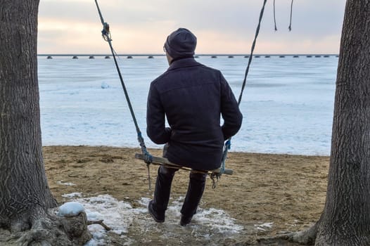 Adult man in a coat swings on a swing and looks thoughtfully at the sea on a cold evening