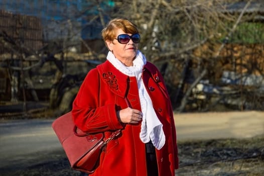 Portrait of stylish 67 year old woman in a bright red coat with a burgundy bag, sunglasses and white scarf on a spring day