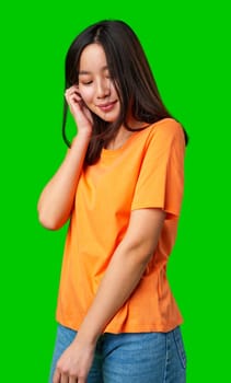 A young woman with long hair stands smiling softly. She wears an orange shirt and blue jeans, creating a vibrant contrast against the bright green backdrop.