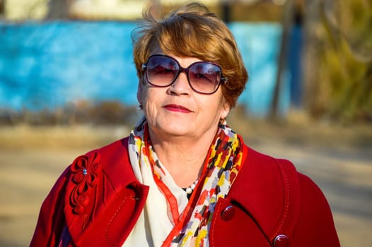 Portrait of stylish 67 year old woman in a bright red coat and sunglasses kindly and sincerely smiling on a spring day