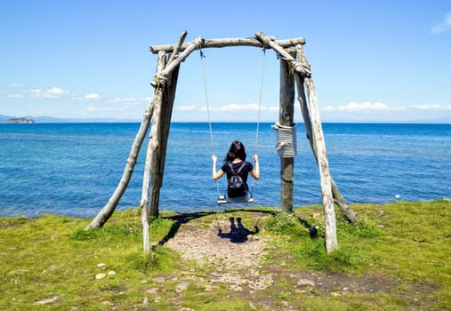 Young brunette woman sitting on wooden swing with her back to the camera on the background of the Japanese sea on a summer day