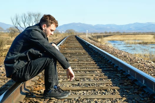 Young man in coat looking down with upset face,sitting on rails. Depressive concept loss of money, loneliness, unemployment. Copy space