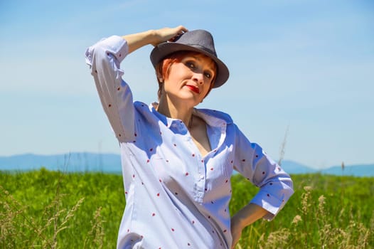 Portrait of Russian red haired woman with bright makeup in hat and  blue blouse on a background of green field