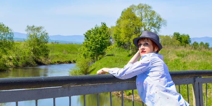 Portrait of Russian red haired woman with bright makeup in hat and  blue blouse on a background of river in countryside. Copy space