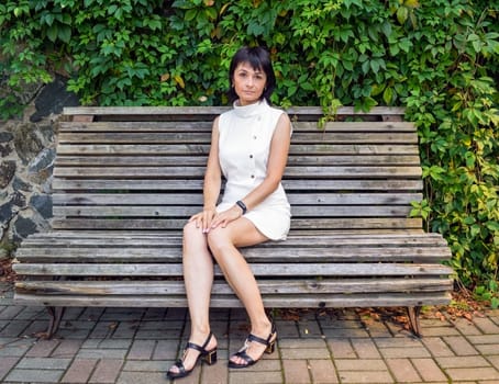 Attractive Russian woman in white sleveless dress sitting on the bench and looking at the camera