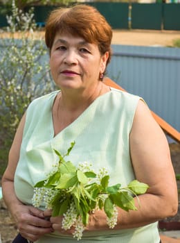 Portrait of 65 y.o. senior woman in sleeveless blouse with 
neutral facial expression sitting and 
holding the spring bouquet of flowering branches of bird cherry tree