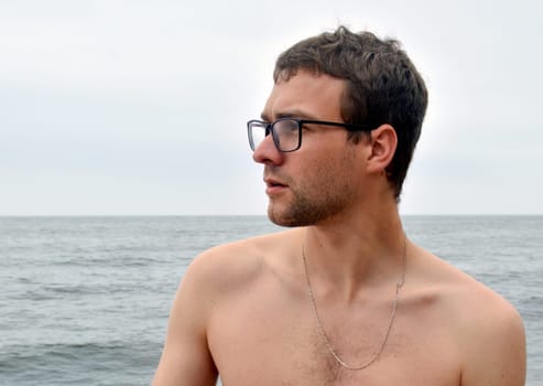 Handsome young guy shirtless, with glasses looking thoughtfully, surprised and attentively to the side against the sea 
in cloudy dark weather on a summer day