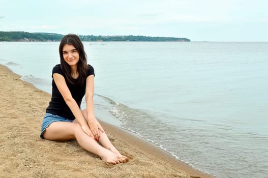 Attractive brunette girl sitting near water on a cloudy summer day. Lake Khanka, Russia.